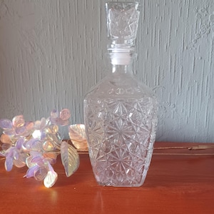 Puede incluir: Una jarra de cristal transparente con un patrón geométrico de estrella. La jarra tiene un tapón cuadrado y probablemente se utiliza para licores o vino. El fondo es una pared verde claro y una superficie de madera.