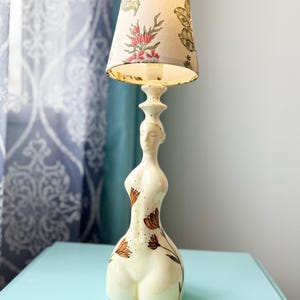 Może przedstawiać: Ceramiczna podstawa lampy w kształcie kobiety z brązowymi wzorami kwiatowymi. Lampa ma biały abażur z kwiatowym wzorem.