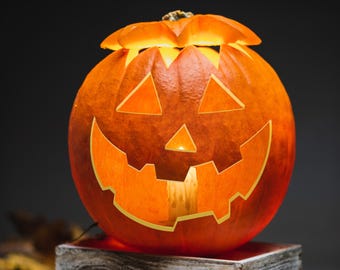 Puede incluir: Una calabaza naranja redonda tallada como jack-o'-lantern con un rostro de ojos triangulares, una nariz pequeña triangular y una boca ancha y dentada. La calabaza está iluminada desde el interior, proyectando un brillo cálido a través de los cortes.