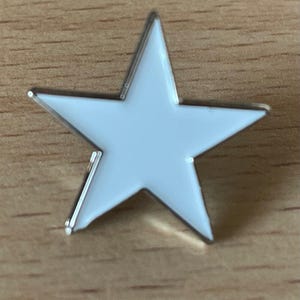 Può includere: Una spilla a forma di stella bianca con un bordo argentato. La spilla è posta su una superficie di legno. La stella ha cinque punte e una finitura liscia e lucida. Questa spilla decorativa può essere utilizzata per accessoriare abiti, borse o altri oggetti.
