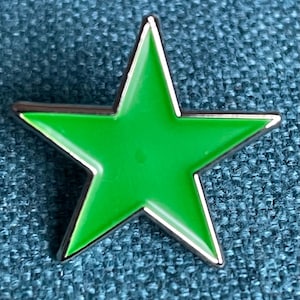 May include: A bright green star-shaped pin with a silver-colored border. The pin is set against a textured blue background. The star has five points and a glossy finish, making it a simple yet eye-catching accessory.