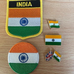 Puede incluir: Una colección de parches y pines con temática de la India. Los parches incluyen un parche rectangular con la palabra "INDIA" y un parche redondo, ambos con las rayas naranja, blancas y verdes de la bandera india y una rueda azul. También hay pines de bandera.