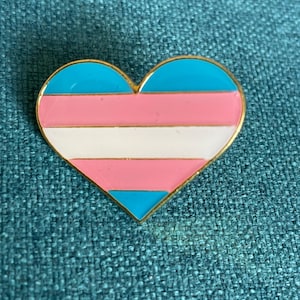 Peut inclure: Une épingle en émail en forme de cœur avec une bordure dorée. L'épingle présente les couleurs du drapeau de la fierté transgenre : bleu clair, rose et blanc. L'épingle est posée sur un fond turquoise texturé.