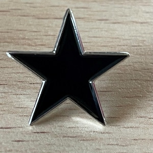 Puede incluir: Un pin negro en forma de estrella con un borde plateado. El pin tiene una superficie brillante y reflectante. La estrella está sobre un fondo de madera clara. Las puntas de la estrella son afiladas y bien definidas.