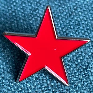 Puede incluir: Un pin de esmalte rojo en forma de estrella con un borde plateado. El pin tiene un acabado brillante y está sobre un fondo azul texturizado. Este accesorio se puede usar para decorar ropa o bolsos.