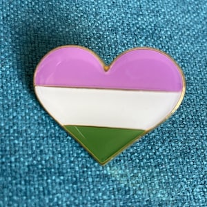 May include: A heart-shaped pin with a gold border. The pin features a design with three horizontal stripes: lavender, white, and green. The pin is set against a textured teal background.