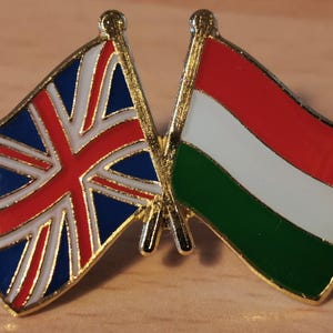 Könnte beinhalten: Goldfarbene Anstecknadel mit zwei gekreuzten Flaggen. Eine ist der Union Jack, mit roten, weißen und blauen Farben. Die andere ist die ungarische Flagge mit roten, weißen und grünen Streifen. Beide Flaggen haben einen goldenen Rand.