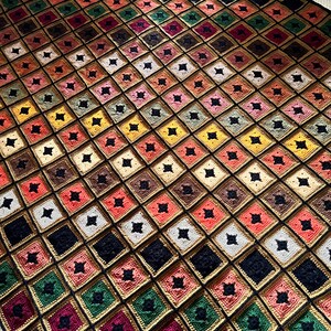 Puede incluir: Una manta de ganchillo hecha a mano con un patrón cuadrado repetitivo. Los cuadrados presentan varios colores, incluyendo negro, blanco, rojo, verde y dorado. La manta está hecha de hilo y tiene una apariencia texturizada, adecuada para la decoración del hogar.