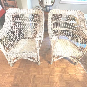 May include: Two matching wicker armchairs. The chairs are light beige with a woven design. The chairs have curved backs and armrests. The chairs are on a parquet floor. The chairs are empty.