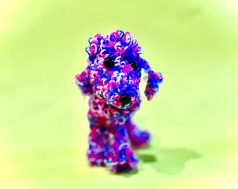 May include: A small, pink, blue, and white rubber band dog figurine. The dog has black eyes and a pink nose.