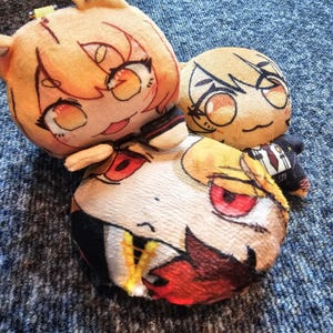 Könnte beinhalten: Drei Plüschtiere mit stilisierten, von Anime inspirierten Gesichtern. Ein großes, rundes Plüschtier hat rote Augen und dunkles Haar, während zwei kleinere Figuren helles Haar und ausdrucksstarke Augen haben. Sie bestehen aus weichem Stoff.