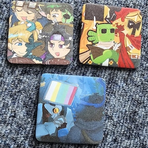 May include: Three square coasters featuring colourful anime-style illustrations. One shows a group of characters, another depicts a green creature and a knight in gold armour, and the third features a character with a television screen for a head.