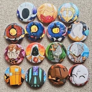 May include: A collection of sixteen circular pin-back buttons featuring diverse illustrated characters and abstract designs in vibrant colors including blue, orange, green, and white, with some buttons displaying a subtle glittery finish.