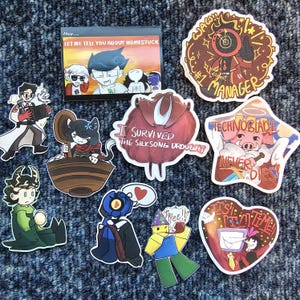 May include: A collection of colorful stickers featuring cartoon characters and text. One sticker reads "LET ME TELL YOU ABOUT HOMESTUCK." Other stickers include "#1 MANAGER," "I SURVIVED THE SILKSONG DROUGHT," and "TECHNOBLADE NEVER DIES!"