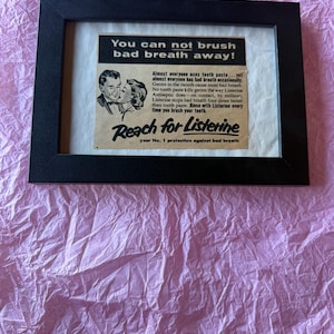 May include: A black rectangular frame containing a vintage advertisement for Listerine. The ad features a black and white illustration of a couple and the text: You can not brush bad breath away! Reach for Listerine, your No. 1 protection against bad breath.