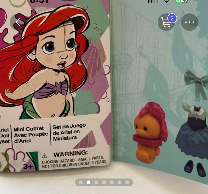 May include: Ariel mini doll set box featuring a cartoon illustration of Ariel with red hair and a purple seashell top. The box is next to a small orange figure with pink hair and a red collar, and a blue dress.