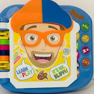 May include: A blue and white interactive toy featuring a cartoon character with orange glasses and a hat. The toy has buttons with icons and the text "Learn & Play" and "It's Me, Blippi!"