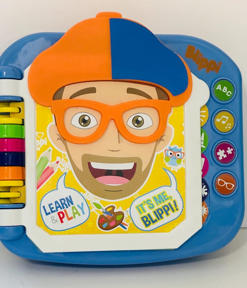 May include: A blue and white interactive toy featuring a cartoon character with orange glasses and a hat. The toy has buttons with icons and the text "Learn & Play" and "It's Me, Blippi!"