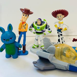 Peut inclure: Ensemble de figurines de personnages de Toy Story, dont Woody, Buzz l'&Eacute;clair, Jessie et un lapin bleu. Un avion jouet est &eacute;galement pr&eacute;sent. Les figurines sont aux couleurs vives et se tiennent sur une surface blanche.