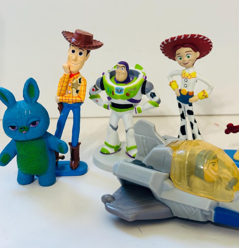 Peut inclure: Ensemble de figurines de personnages de Toy Story, dont Woody, Buzz l'&Eacute;clair, Jessie et un lapin bleu. Un avion jouet est &eacute;galement pr&eacute;sent. Les figurines sont aux couleurs vives et se tiennent sur une surface blanche.