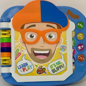 May include: A blue and white children's learning toy featuring a cartoon character with orange glasses and a hat. The toy has colorful buttons and the text "Learn & Play" and "It's Me, Blippi!"