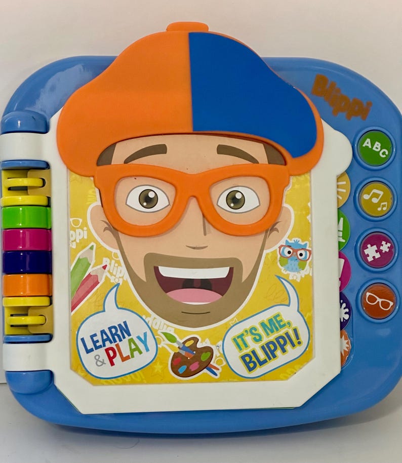May include: A blue and white children's learning toy featuring a cartoon character with orange glasses and a hat. The toy has colorful buttons and the text "Learn & Play" and "It's Me, Blippi!"