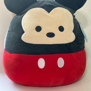 Op de afbeelding: Een pluche Mickey Mouse kussen in een afgeronde vorm. De bovenste helft is zwart met grote, ronde oren en een cr&egrave;mekleurig gezicht. De onderste helft is rood met twee witte, ovaalvormige knopen.