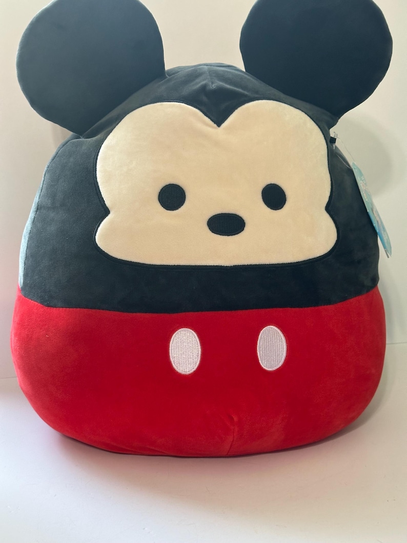 Op de afbeelding: Een pluche Mickey Mouse kussen in een afgeronde vorm. De bovenste helft is zwart met grote, ronde oren en een cr&egrave;mekleurig gezicht. De onderste helft is rood met twee witte, ovaalvormige knopen.