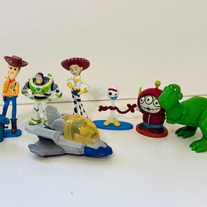 Peut inclure: Une collection de figurines color&eacute;es de personnages de Toy Story, dont Woody, Buzz l'&Eacute;clair, Jessie et un dinosaure vert. Les figurines sont en plastique et sont pos&eacute;es sur une surface blanche.