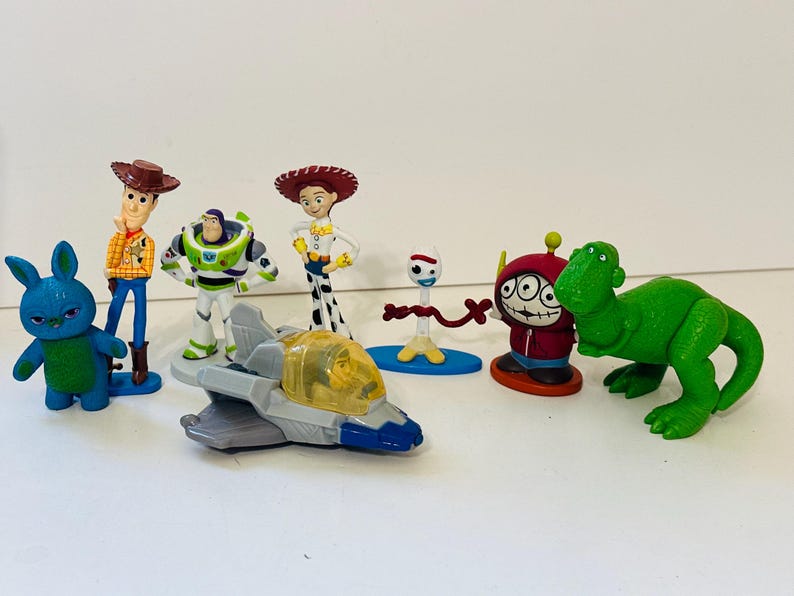 Peut inclure: Une collection de figurines color&eacute;es de personnages de Toy Story, dont Woody, Buzz l'&Eacute;clair, Jessie et un dinosaure vert. Les figurines sont en plastique et sont pos&eacute;es sur une surface blanche.