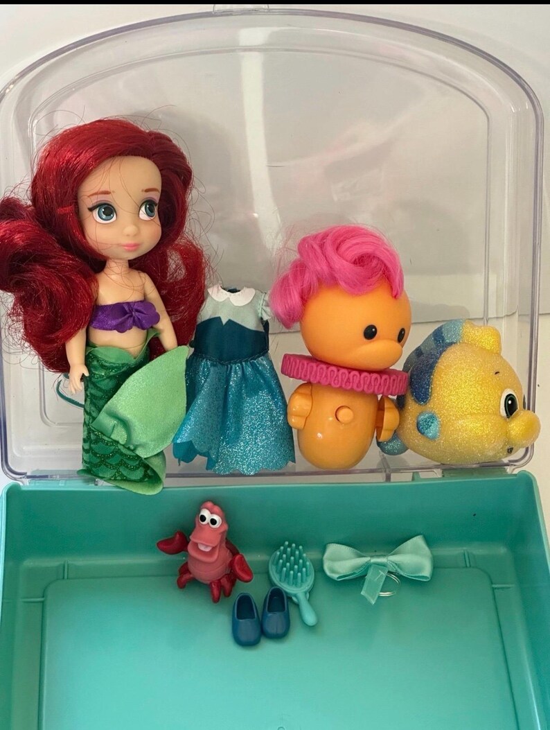May include: Ariel doll with red hair, a purple top, and a green tail, along with other character figures and accessories. Includes a blue dress, a pink-haired figure, a yellow fish, a red crab, and a hairbrush, all inside a clear plastic case.