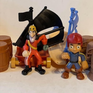 May include: A toy pirate ship with two pirate figures and several brown barrels. The ship has a black sail and a red base. One pirate figure is wearing a red outfit and the other is wearing a blue outfit.
