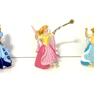 May include: Three miniature figurines of princesses in colorful gowns. The first princess wears a blue and navy dress and holds a blue bird. The second princess is in a pink and yellow dress. The third princess is in a blue and yellow dress and holds a wand.