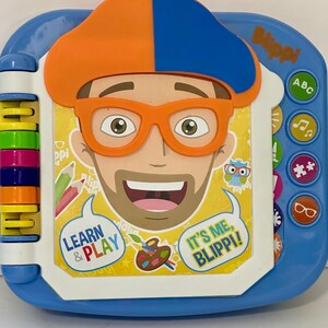 May include: A blue and white children's learning toy featuring a cartoon character with orange glasses and a hat. The toy has buttons with icons and the text "Learn & Play" and "It's Me, Blippi!"