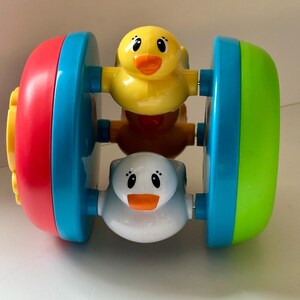 May include: A colorful baby toy featuring a rolling design with three duck figures. The toy has red, blue, and green accents. The ducks are yellow, white, and brown.