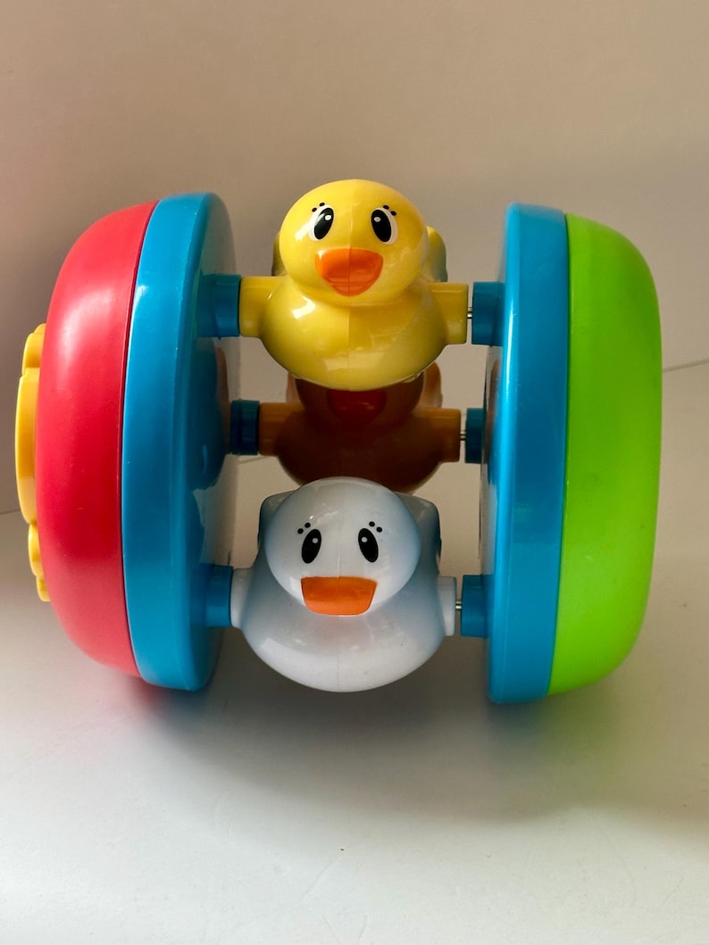 May include: A colorful baby toy featuring a rolling design with three duck figures. The toy has red, blue, and green accents. The ducks are yellow, white, and brown.