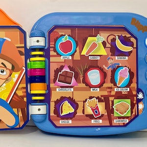 May include: A blue and orange Blippi learning toy with colorful buttons and illustrations of food items like an apple, carrot, and ice cream. The toy has a flip-open design and features a cartoon character.