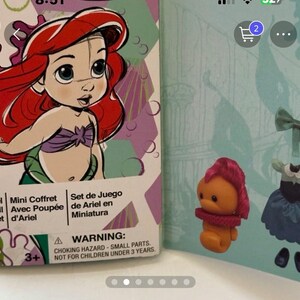May include: Ariel mini doll set box featuring a cartoon illustration of Ariel with red hair and a purple top. The box is next to a small orange figure with pink hair and a red collar, and a blue dress.