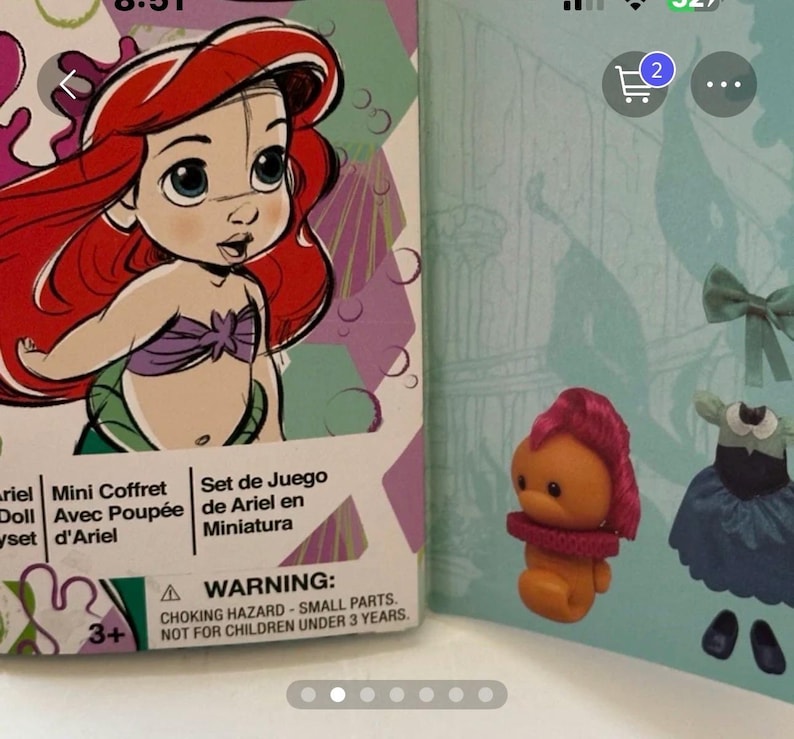 May include: Ariel mini doll set box featuring a cartoon illustration of Ariel with red hair and a purple top. The box is next to a small orange figure with pink hair and a red collar, and a blue dress.