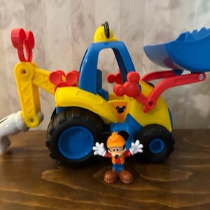 May include: A colorful toy construction vehicle with a blue scoop and cab, yellow body, and red accents. A Mickey Mouse figurine in a construction outfit stands in front of the vehicle. The toy has black tires and is designed for children.