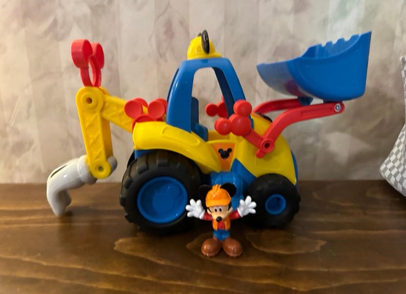 May include: A colorful toy construction vehicle with a blue scoop and cab, yellow body, and red accents. A Mickey Mouse figurine in a construction outfit stands in front of the vehicle. The toy has black tires and is designed for children.