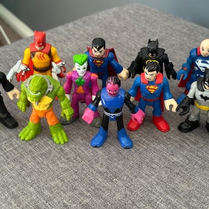 May include: A collection of colorful superhero action figures, including Batman, Superman, and The Flash. The figures are made of plastic and feature detailed designs. The characters are posed in various action stances.