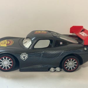 May include: A black toy race car with a carbon fiber pattern, featuring the number 95 on the roof and a large red rear spoiler. The car has white wheels with red rims, dual side exhaust pipes, and a Piston Cup logo on the hood and side panel.