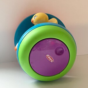 May include: A Playskool toy with a yellow duck on top. The toy is a combination of bright colors, including teal, green, and purple. The Playskool logo is visible on the purple surface. The toy is designed for young children.