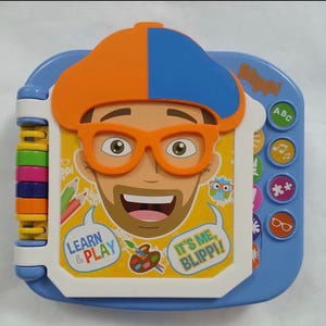 May include: A blue and white children's learning toy featuring a cartoon character with orange glasses and a hat. The toy has buttons with ABCs, music notes, and other symbols. Text on the toy reads "Learn & Play" and "It's Me, Blippi!"