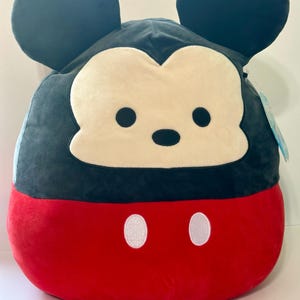 Op de afbeelding: Een Mickey Mouse pluche kussen met een zwarte kop en oren, een cr&egrave;mekleurig gezicht en een rode body. Het kussen heeft twee witte ovale knopen op het rode gedeelte. Het ontwerp doet denken aan het iconische Disney-personage.