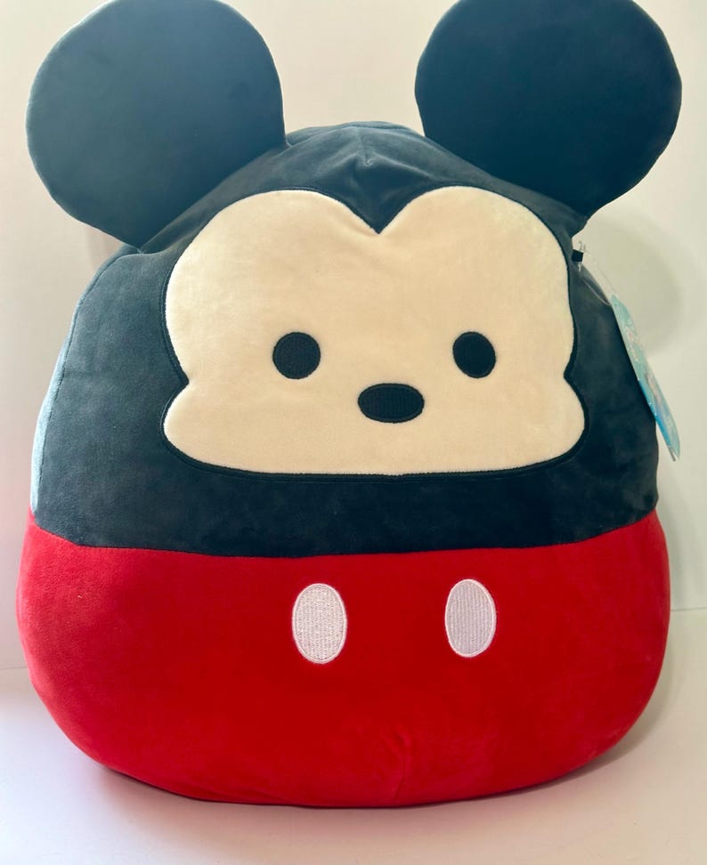 Op de afbeelding: Een Mickey Mouse pluche kussen met een zwarte kop en oren, een cr&egrave;mekleurig gezicht en een rode body. Het kussen heeft twee witte ovale knopen op het rode gedeelte. Het ontwerp doet denken aan het iconische Disney-personage.