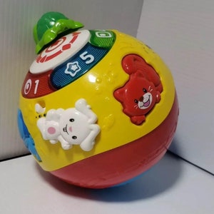 May include: A colorful, interactive baby toy ball with a green top. The ball features a red, yellow, and blue design with animal shapes and numbers. The toy has a white bunny, a red cat, and the number 5.