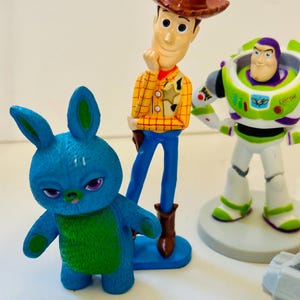 Peut inclure: Figurines de personnages de Toy Story, dont Woody avec un chapeau de cow-boy, Buzz l'&Eacute;clair dans une combinaison spatiale verte et blanche, et un personnage bleu en forme de lapin. Les figurines sont color&eacute;es.