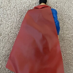 May include: A Superman action figure viewed from the back, showcasing a red cape and blue suit. The figure has black hair and a red collar. The cape is a deep red color.